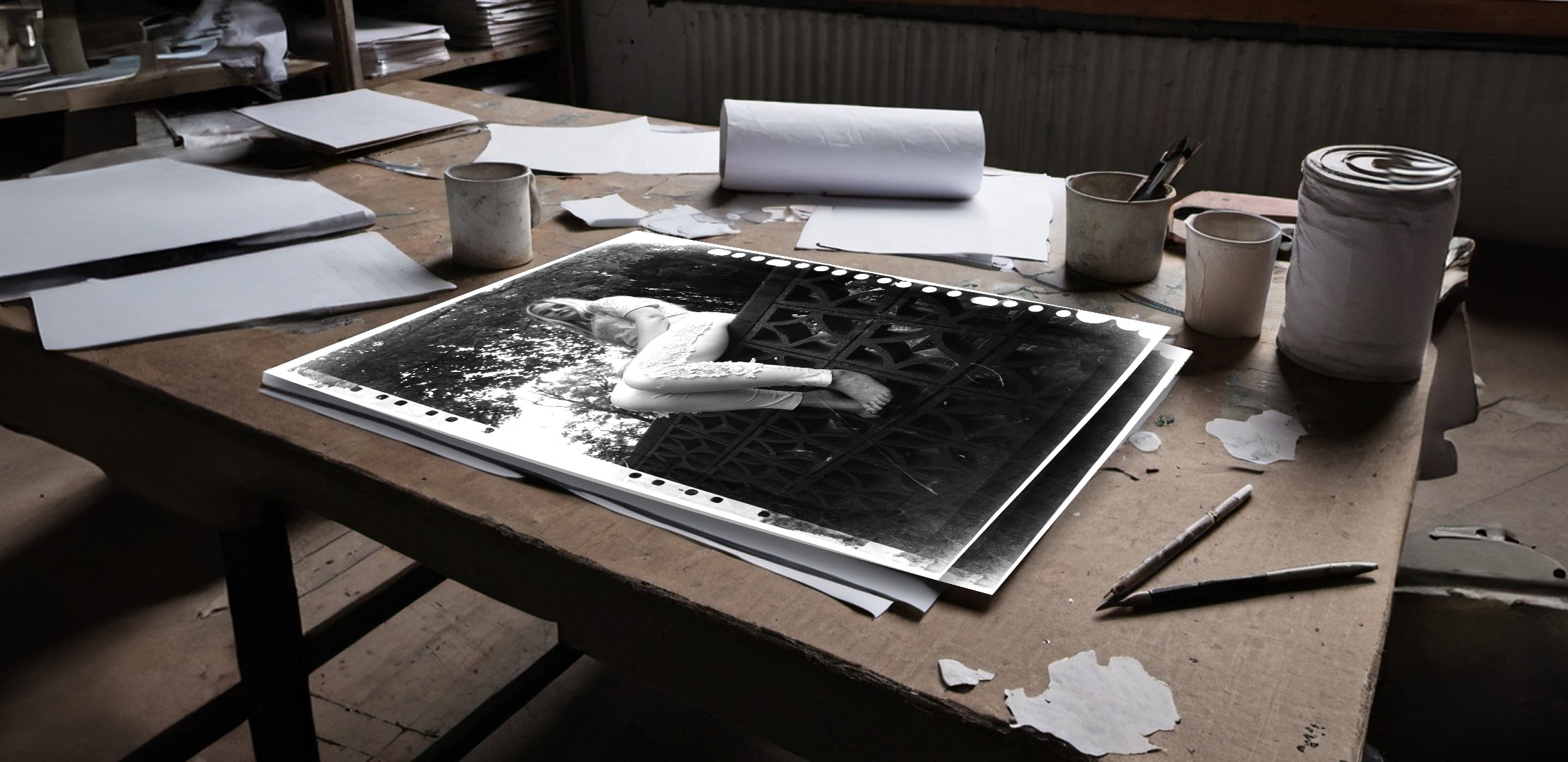 Black and white photograph of a woman on a rustic worktable surrounded by art supplies and papers.